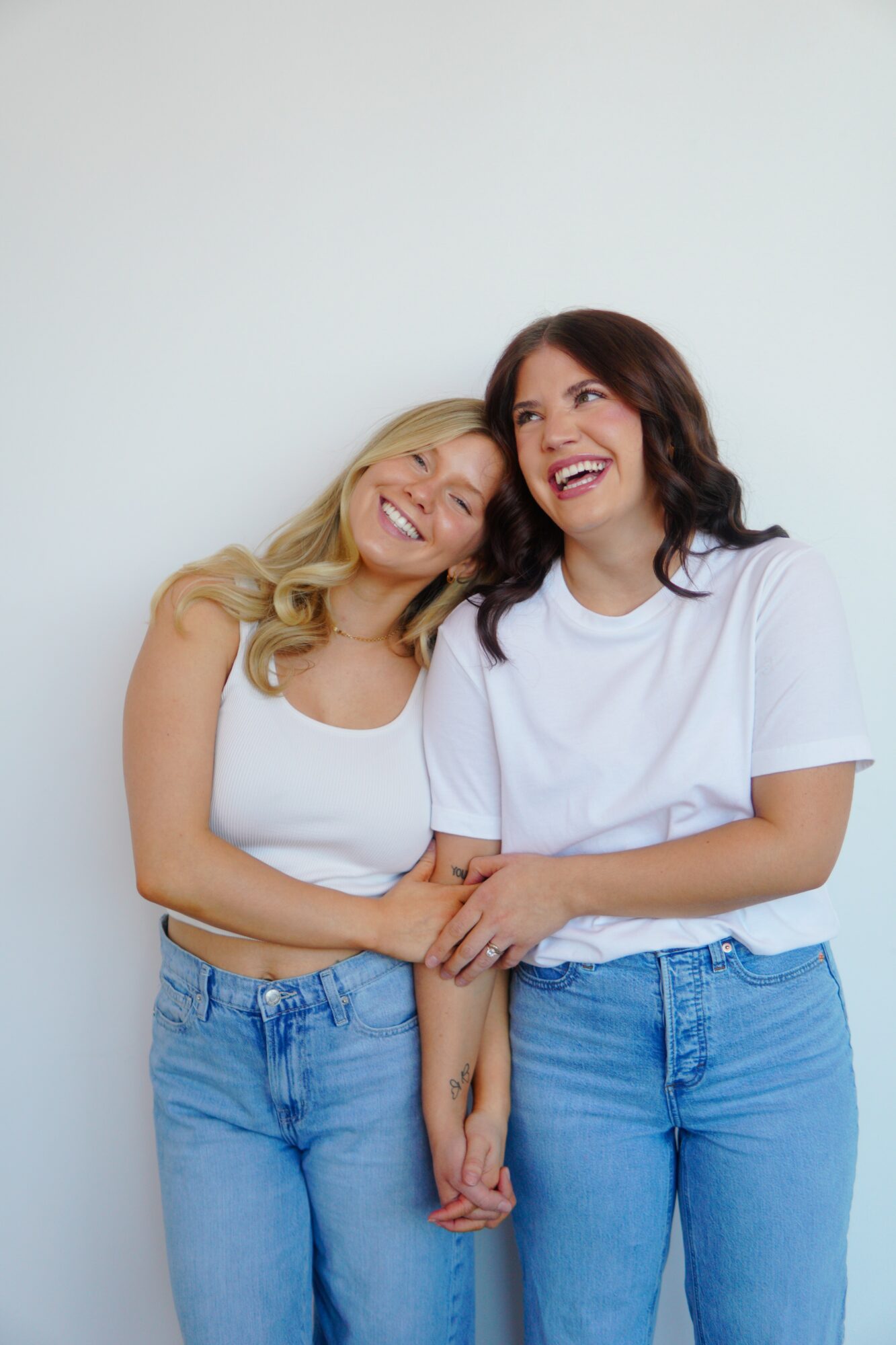 Two women smiling and holding hands, standing close together against a plain light background.