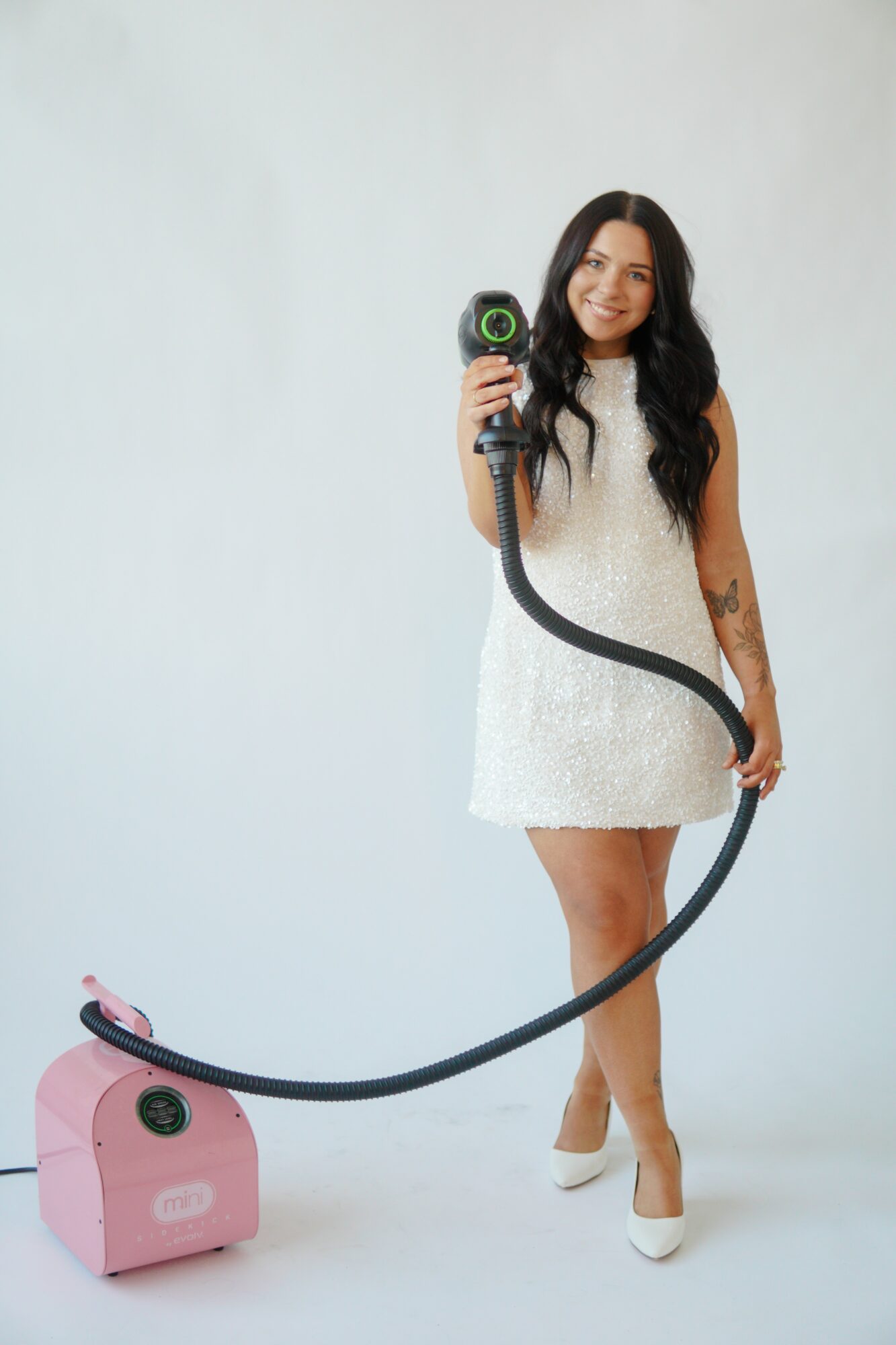 Woman in white dress holding a vacuum cleaner hose, smiling, standing on a white background.