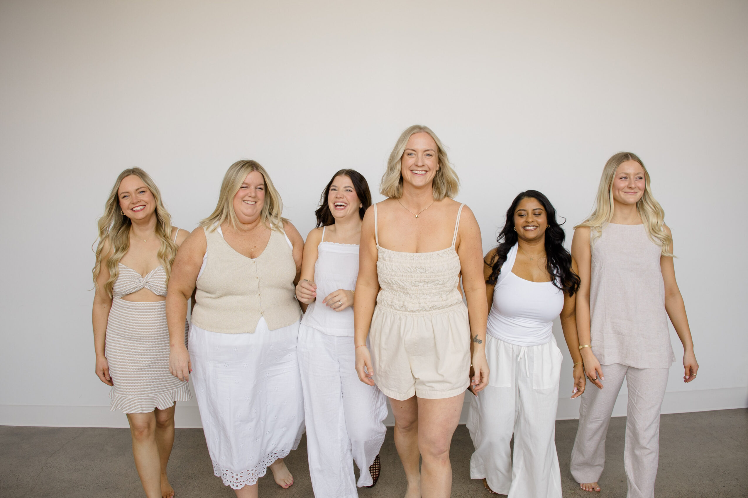 Six women standing in a row against a plain wall, smiling and looking at the camera.