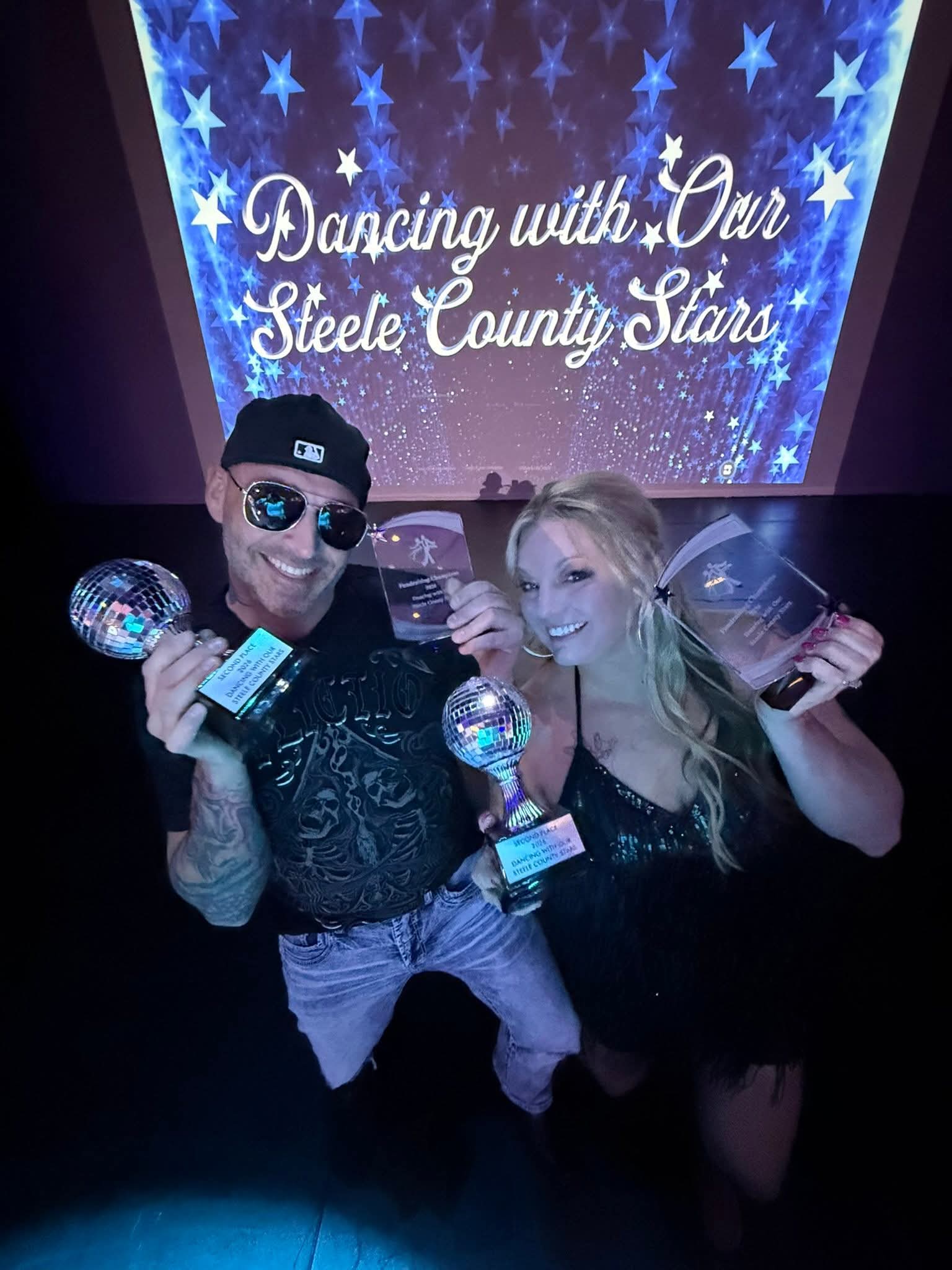 Two people holding trophies and a disco ball, smiling in front of a starry backdrop with text about dancing and stars.