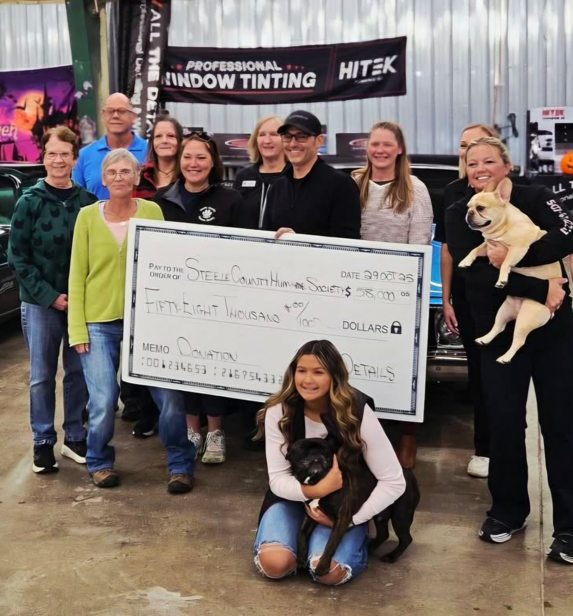 Group of people holding a large check, some smiling, with a woman holding a dog, in an indoor setting.