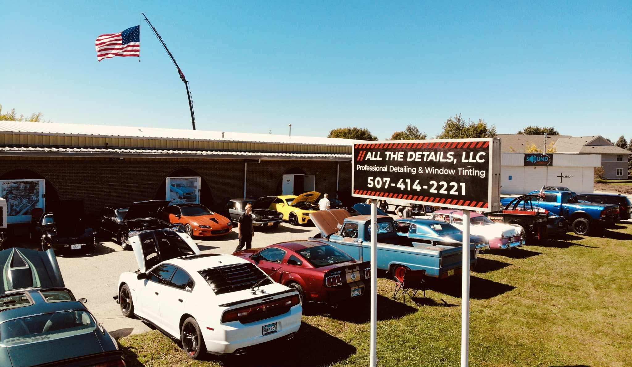 Car dealership lot with various cars, a building, and an American flag on a pole, under a blue sky.