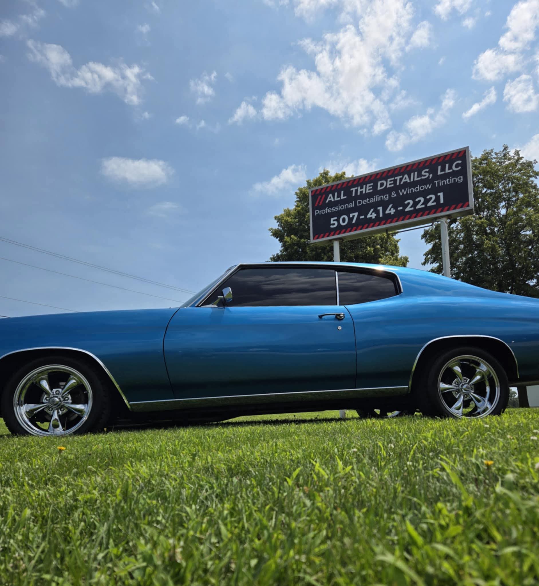 Blue vintage car parked on grass with a sign and trees in the background under a partly cloudy sky.