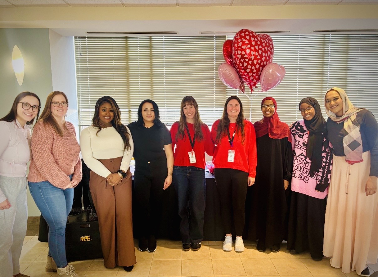 Group of nine women standing indoors with a heart-shaped balloon decoration above them.