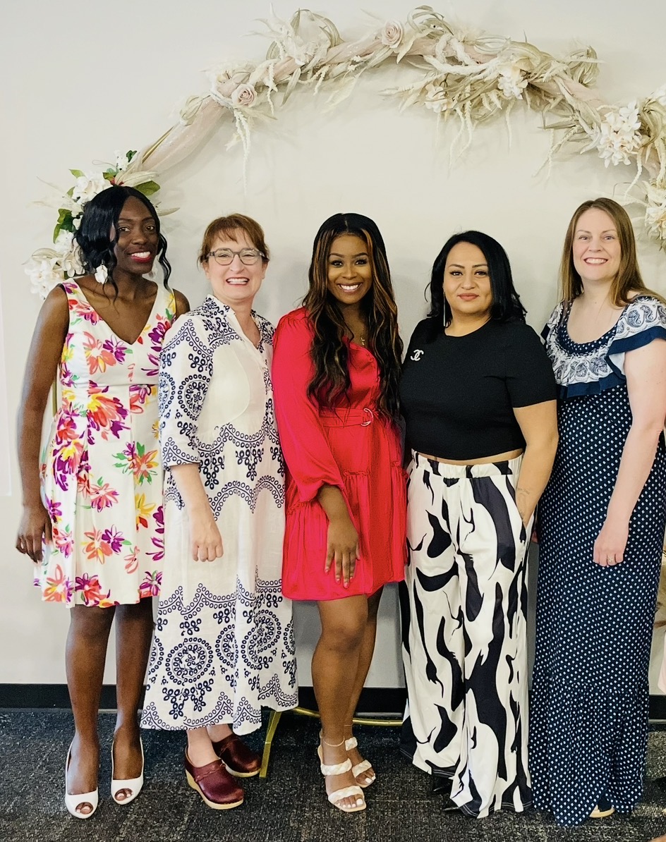 Five women standing together in front of a floral arch, smiling, wearing colorful dresses and pants.