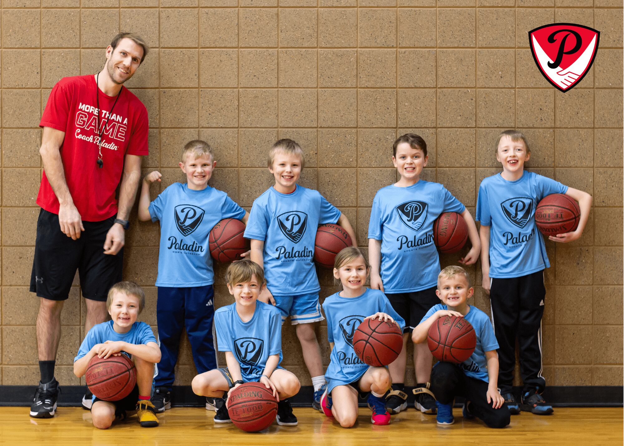 Group of young boys and a coach in basketball uniforms posing with basketballs against a brick wall.