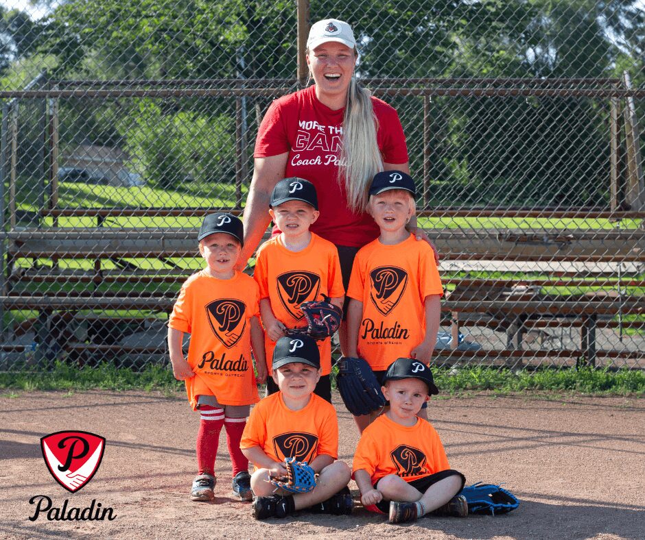 A woman with long blonde hair and a white cap stands behind four children in baseball uniforms on a field.