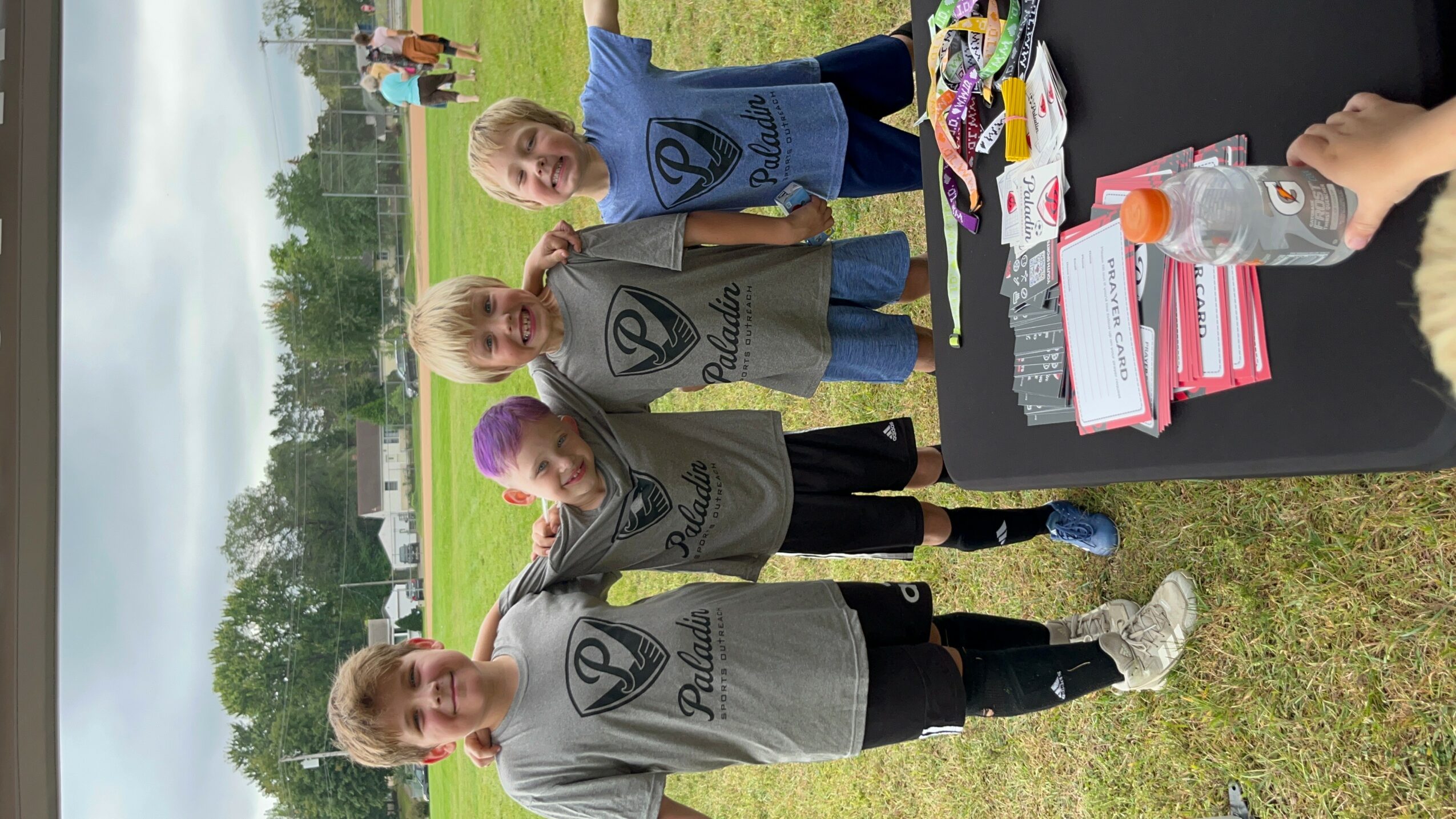 Four children standing outdoors near a table with food and drinks, smiling at the camera.