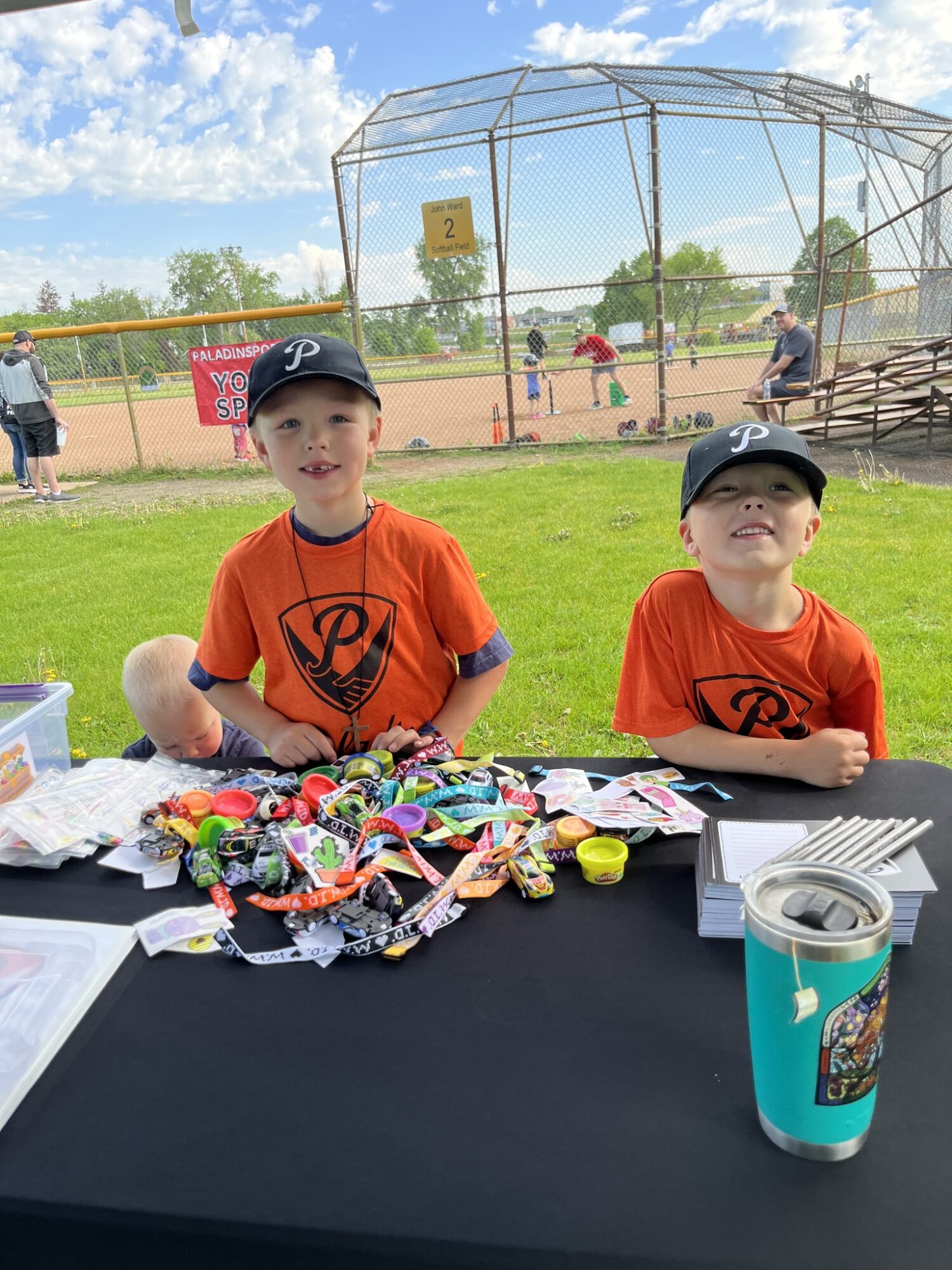 Two children in orange shirts and black caps sit behind a table with candy and a drink cup, baseball field in background.