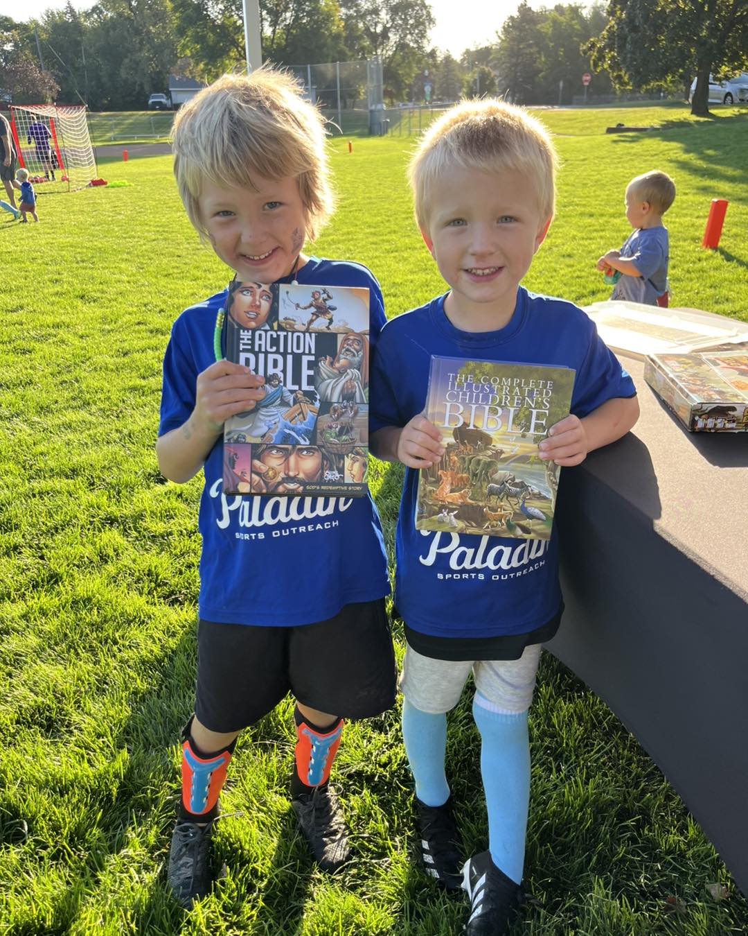 Two children standing outdoors on grass, holding books and smiling, with trees and a person in the background.
