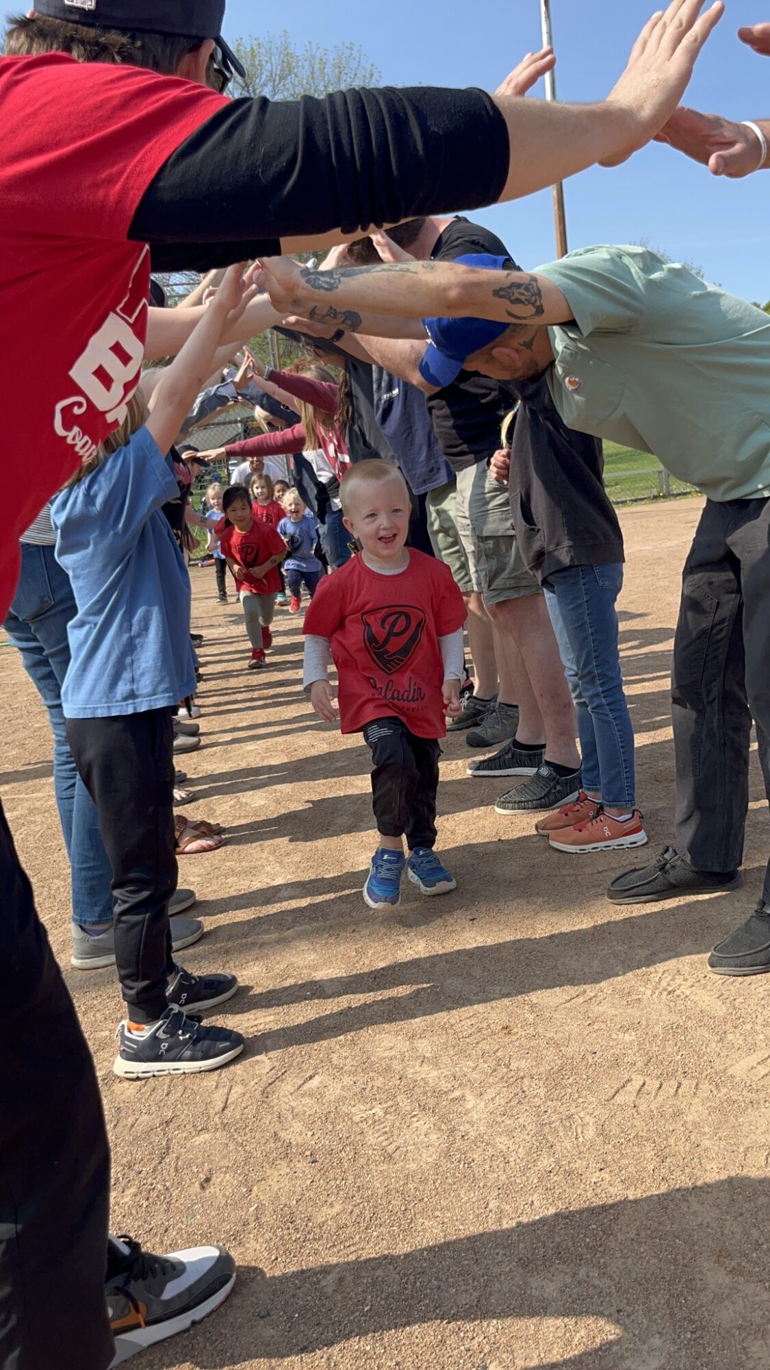 Children and adults form a tunnel with arms for a child to walk through outdoors.