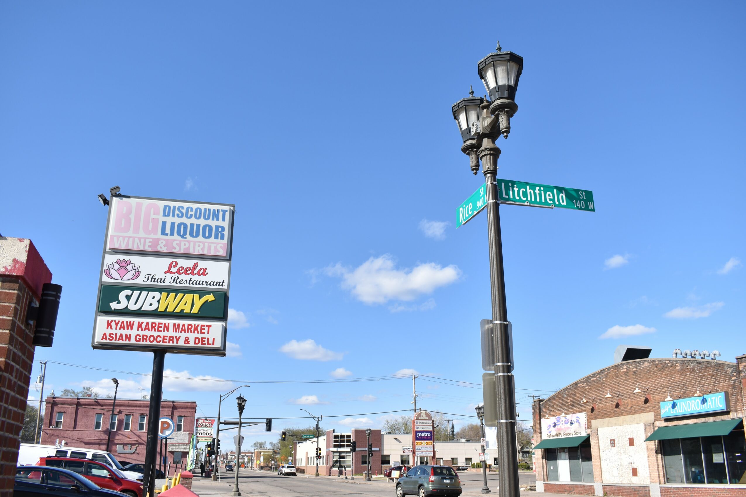 Street corner with street signs, lamp post, and storefronts under a blue sky with clouds.