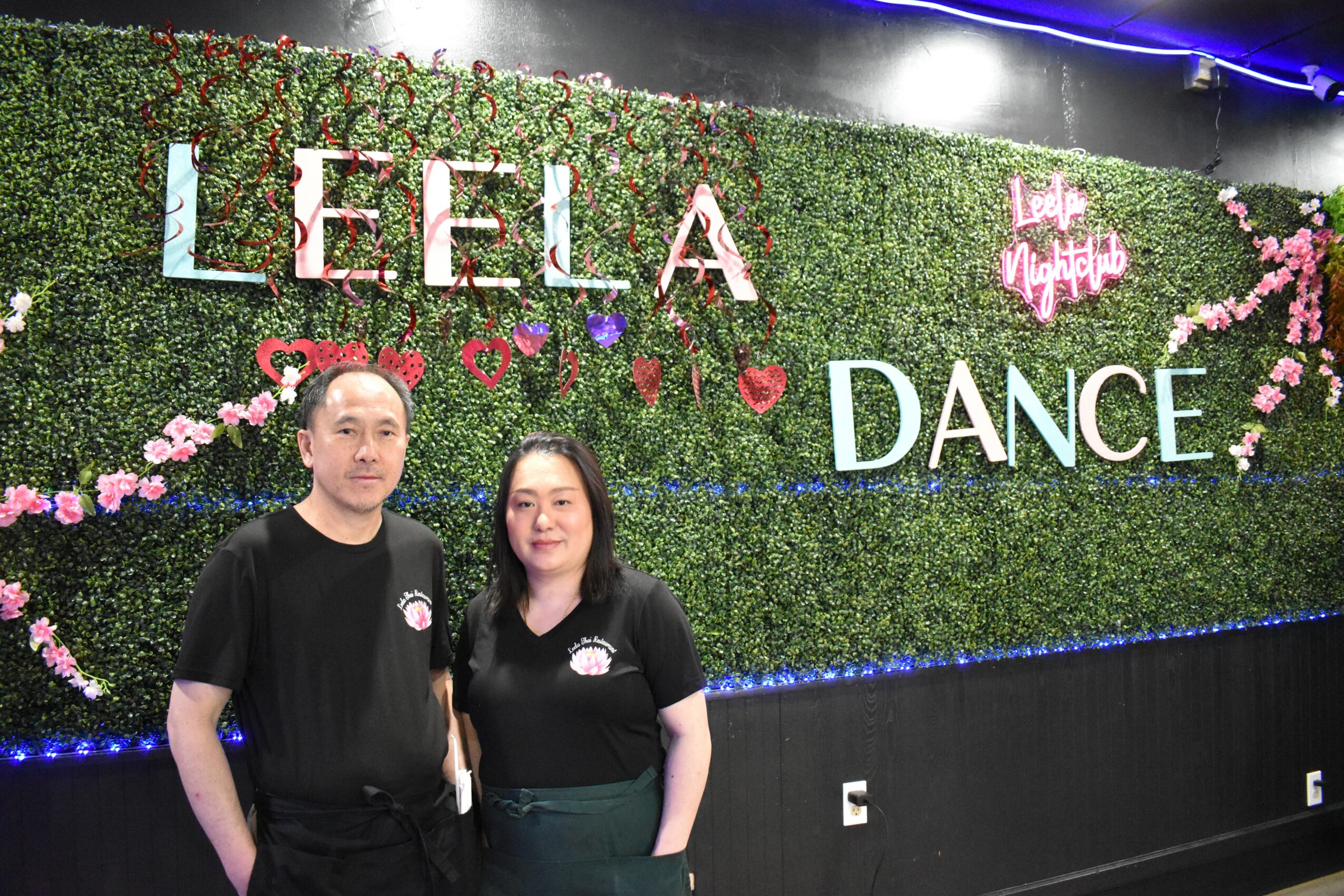 Two people stand in front of a decorated wall with the words 'LEELA' and 'DANCE' and floral decorations.