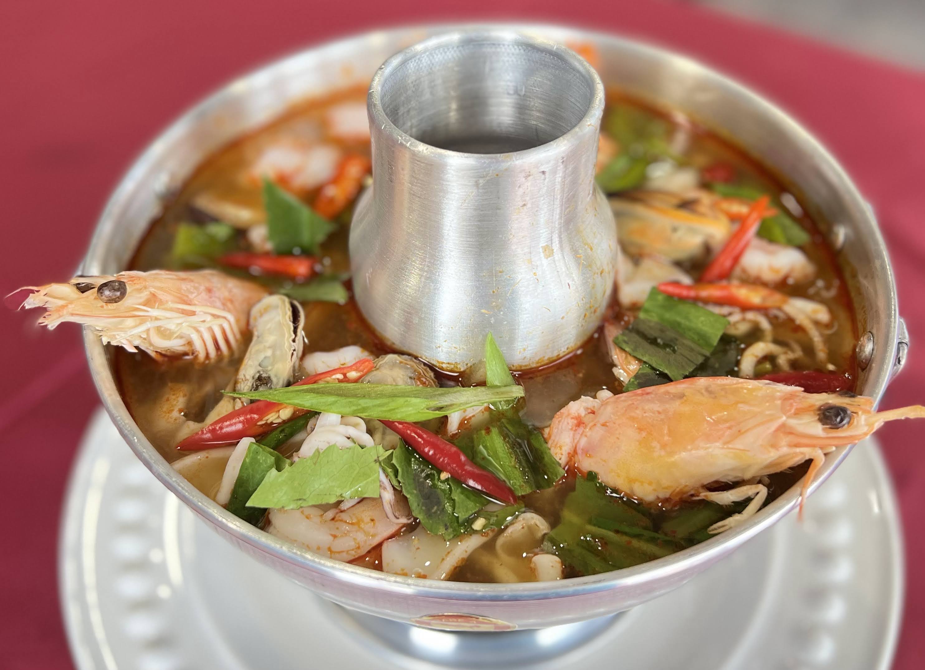 Metal bowl filled with spicy soup containing chicken, vegetables, and red chili peppers, on a white plate with a red background.