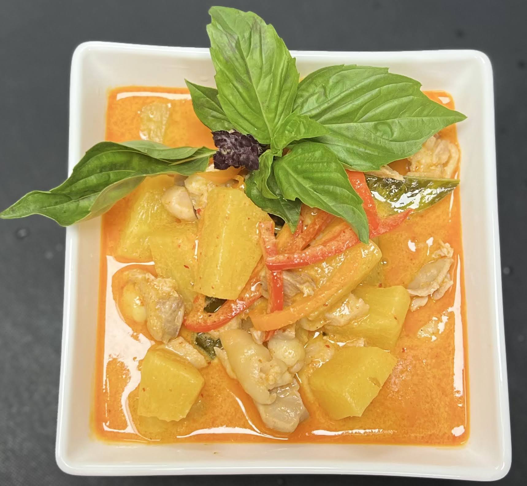 Square bowl with orange-colored soup, topped with basil leaves and garnished with vegetables and chicken pieces.