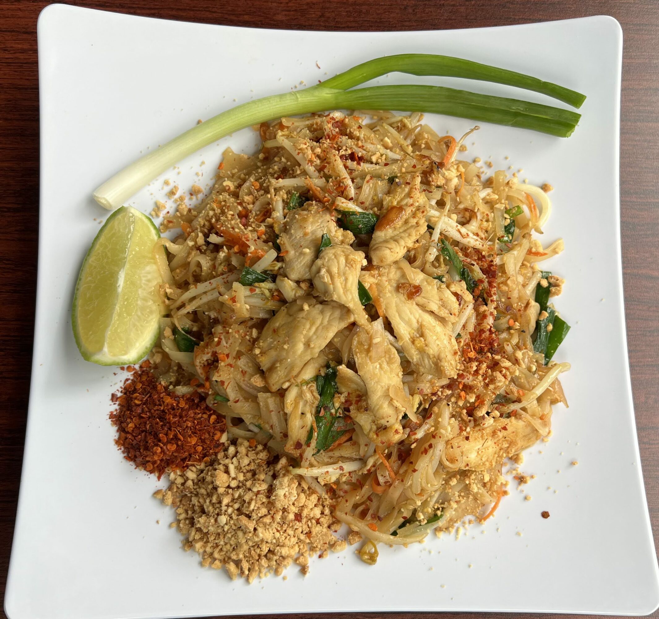Plate of stir-fried noodles with vegetables, lime wedge, green onions, and crushed peanuts on a white square plate.