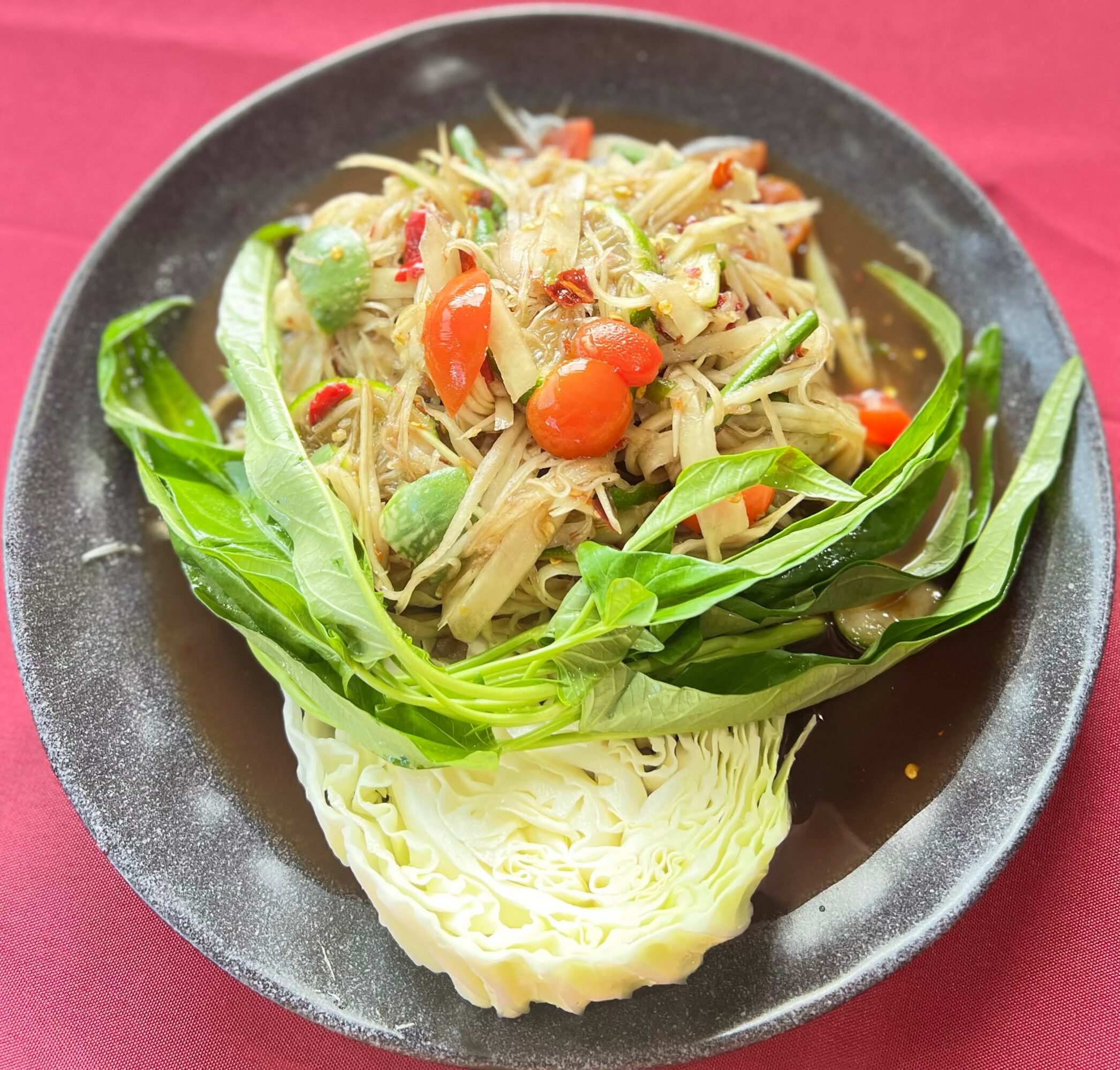 Noodle salad with vegetables on a black plate, garnished with leafy greens, served on a red table.