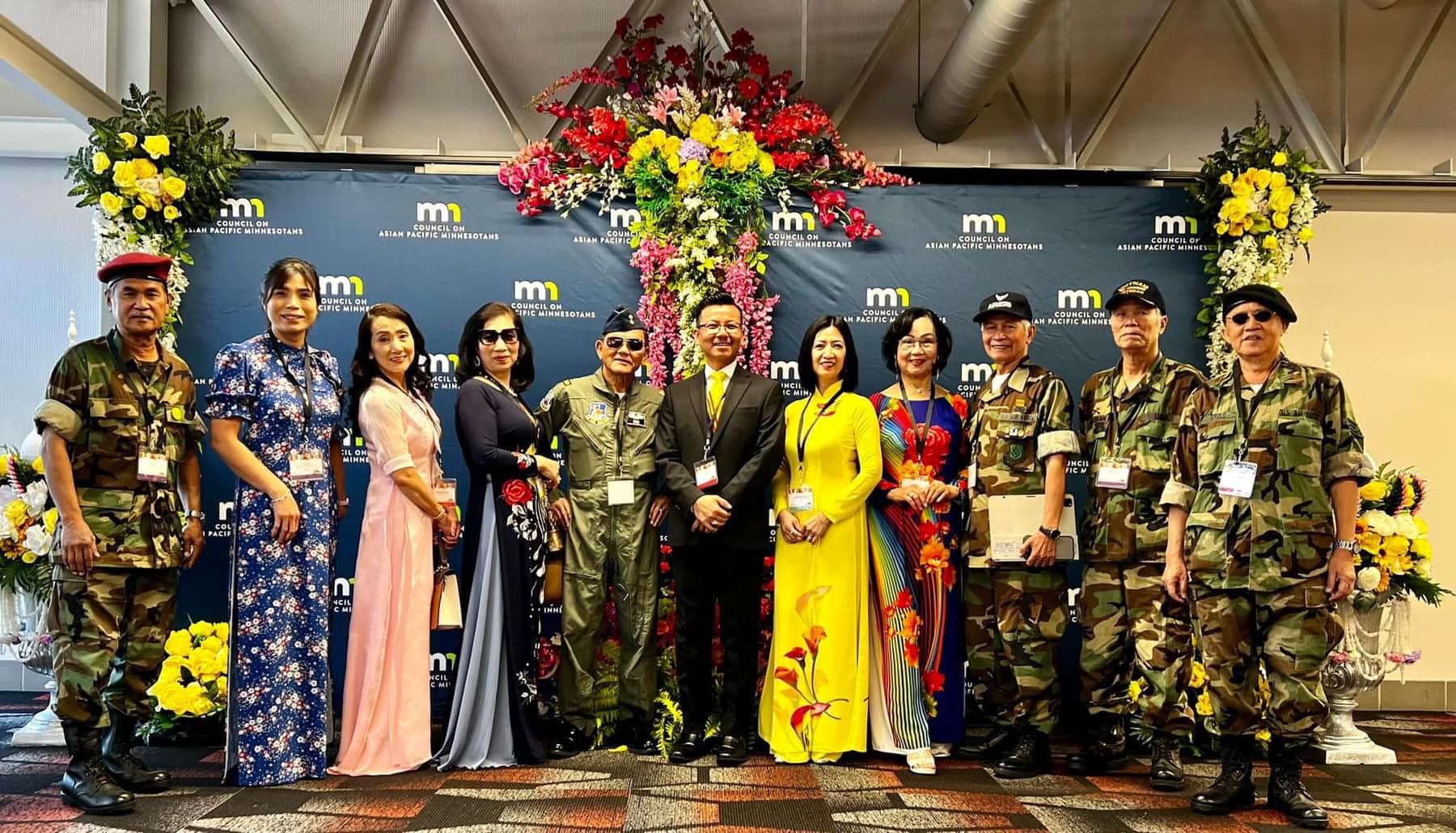 Group of ten people standing in front of a floral display and backdrop with logos, some in military uniforms and traditional attire.