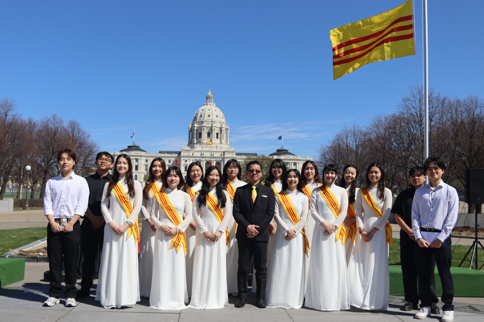Group of people in white dresses with yellow sashes standing outdoors in front of a government building, with a flag flying nearby.