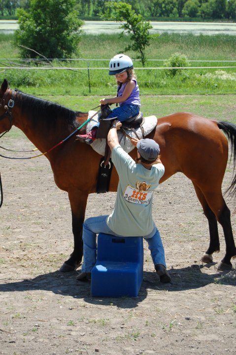 Child wearing helmet riding a horse with an instructor adjusting the saddle outdoors.
