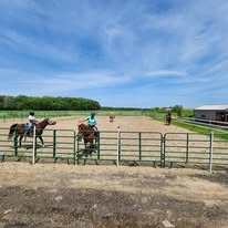 People riding horses in an outdoor riding arena with a fence, under a blue sky with clouds.