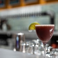 A cocktail glass with a dark red drink and a lime wedge garnish, blurred bar background.