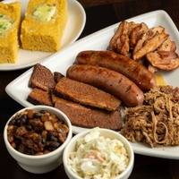 Plate with sausages, sliced meat, pulled pork, baked beans, coleslaw, and cornbread pieces.