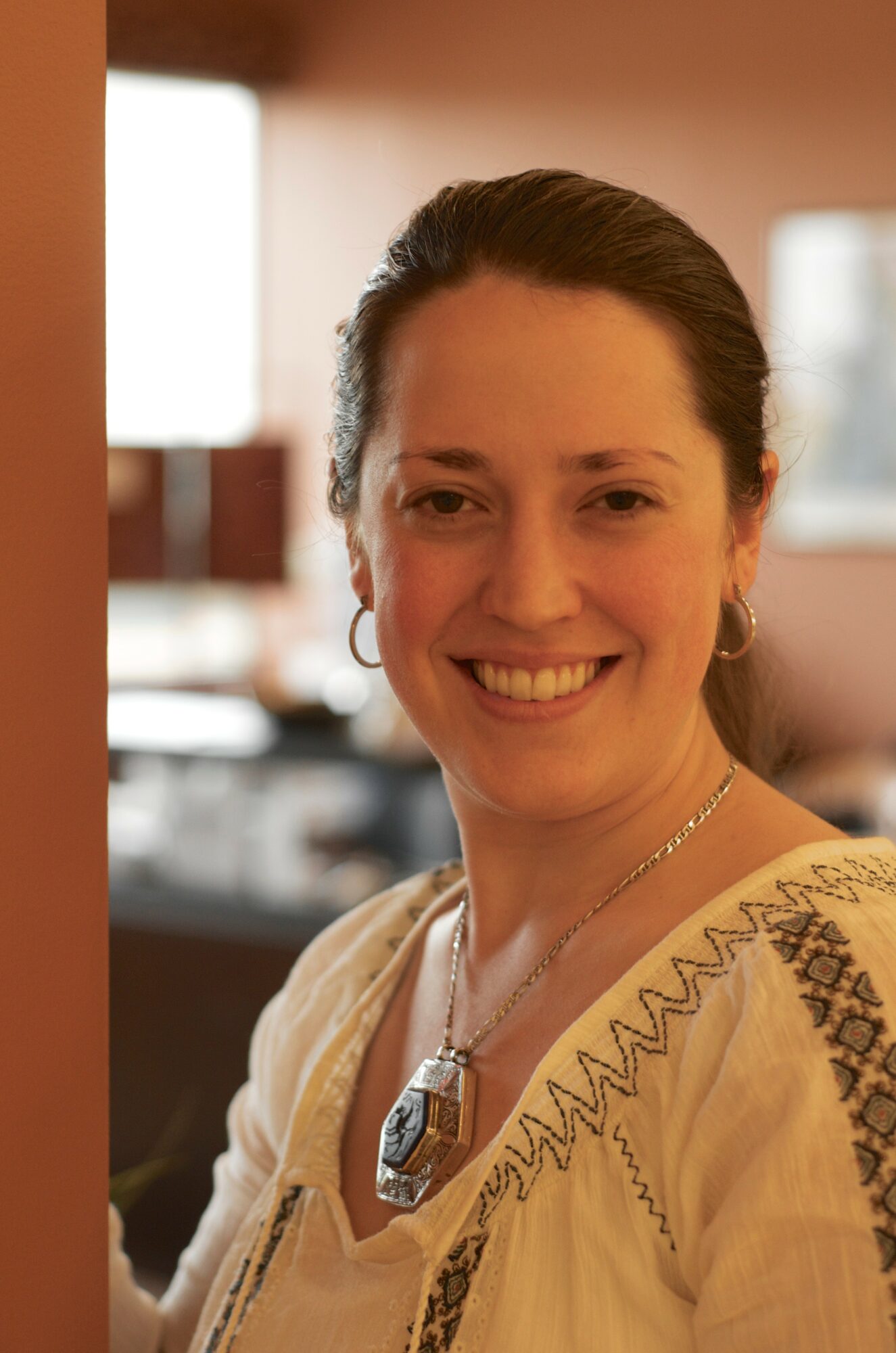 Smiling woman with dark hair, wearing earrings and a necklace, indoors with blurred background.