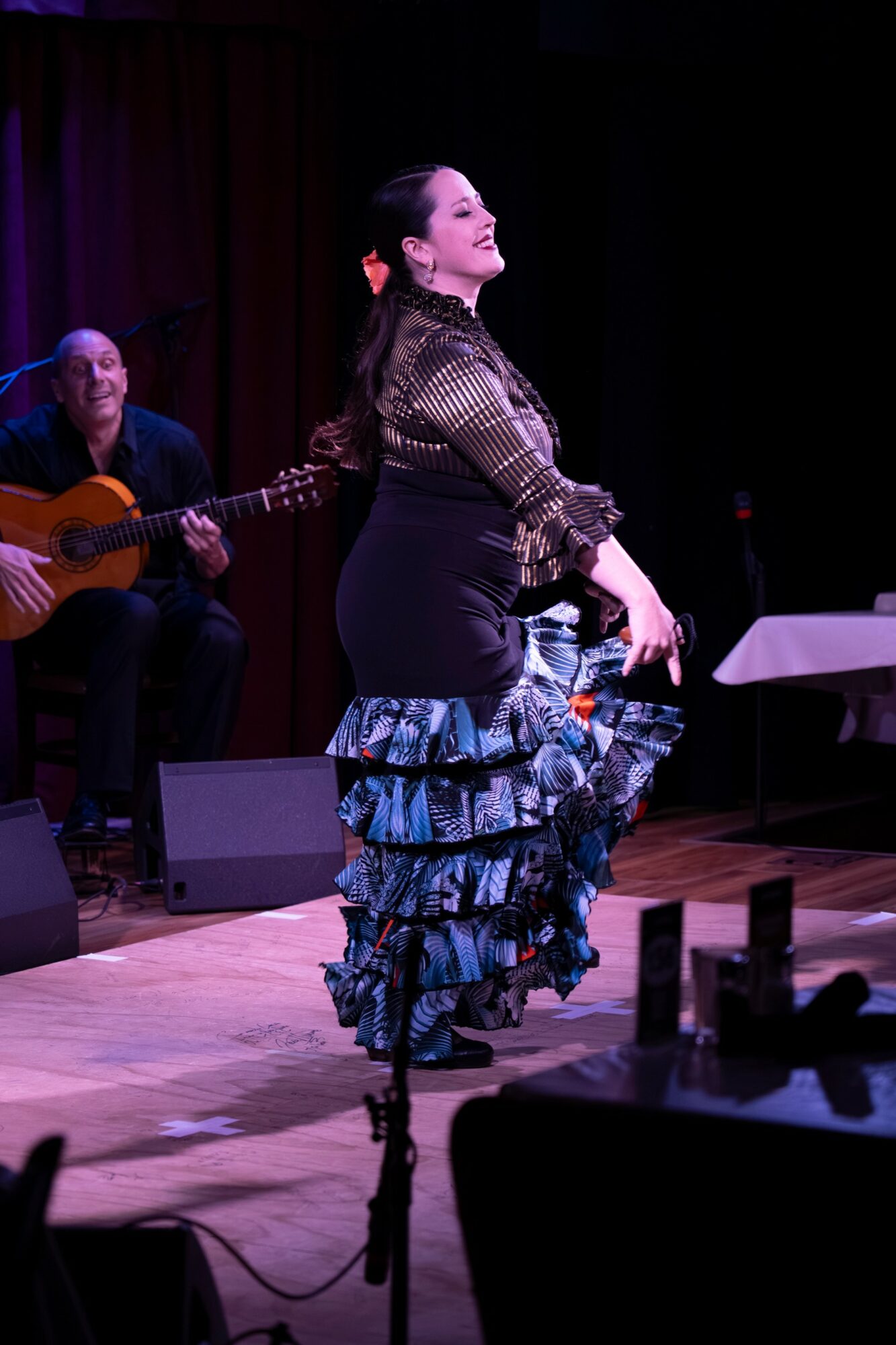 A woman dances on stage with a man playing guitar in the background, stage lighting highlights her movements.