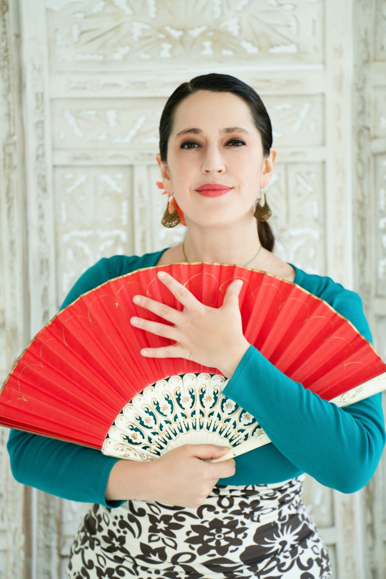 Woman holding a red fan in front of a white textured background, wearing a teal top and earrings.