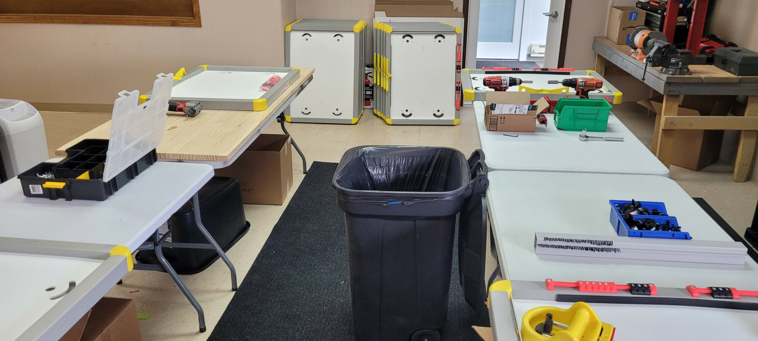 Room with tables, boxes, and a black trash bin in the center, workspace with tools and supplies on tables.
