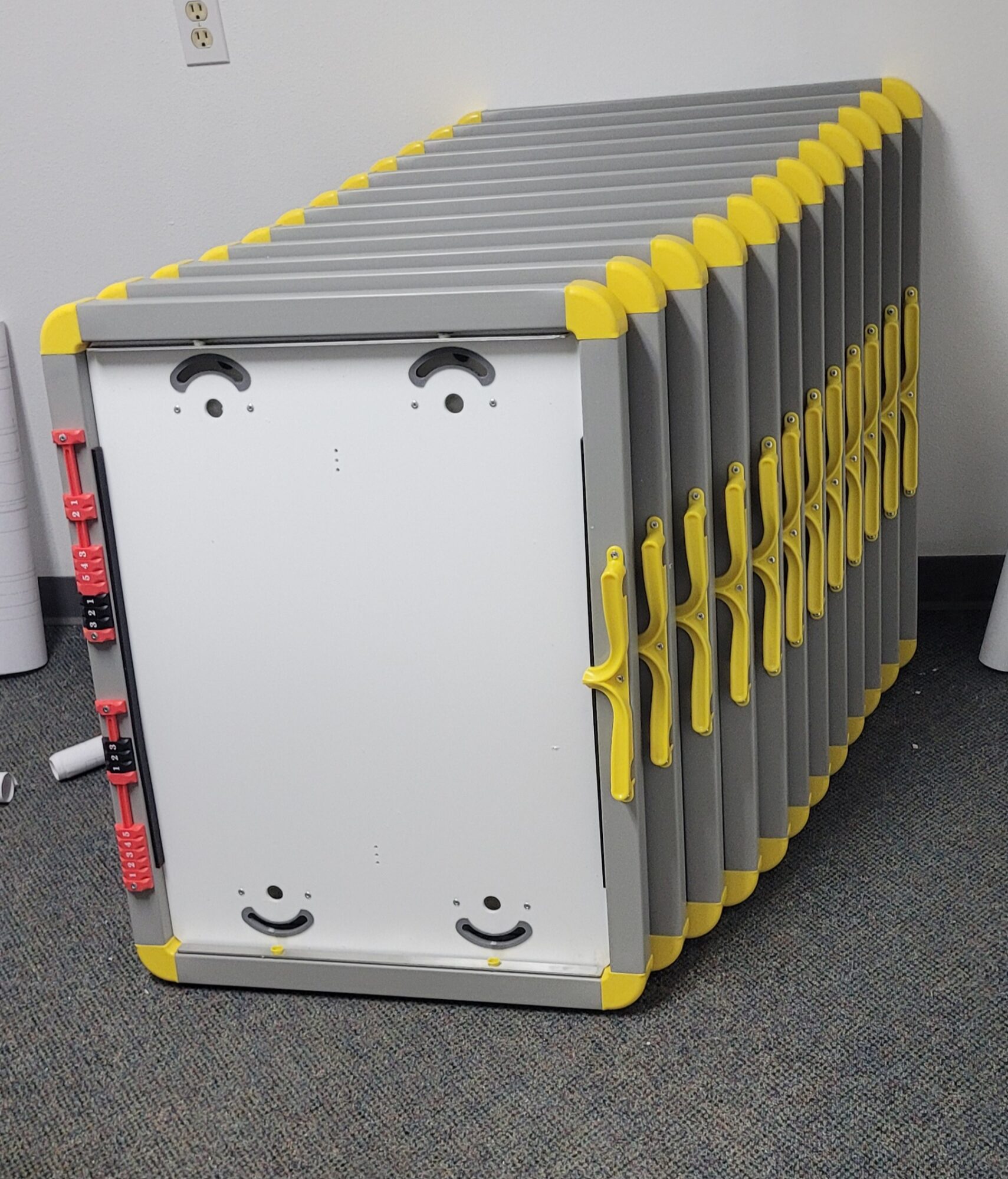 Stack of gray folding tables with yellow edges, stored against a wall in a room.