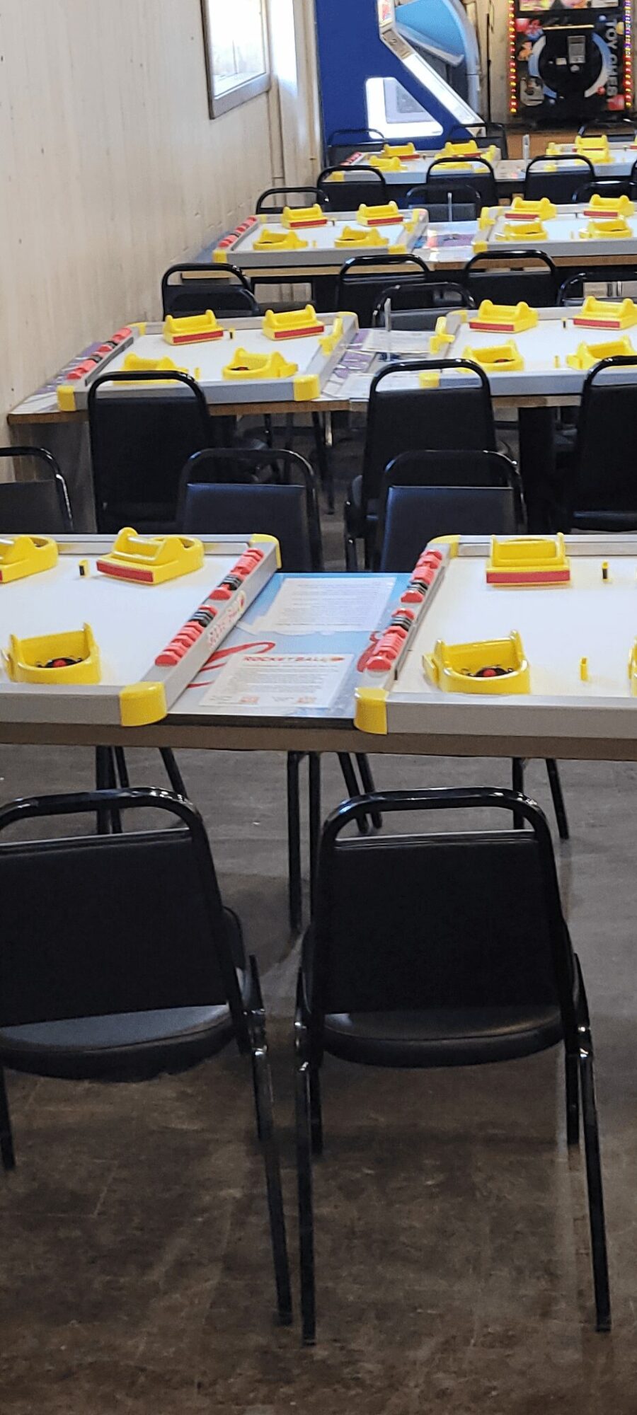 Empty tables with yellow and white items, black chairs, and a gaming machine in the background.