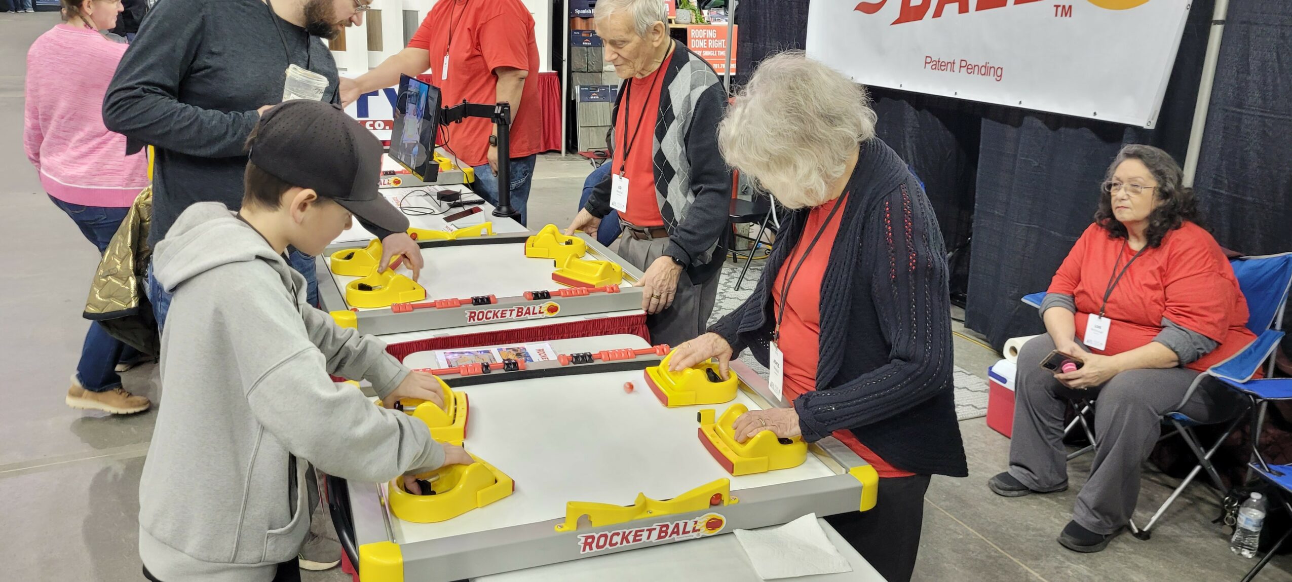 People playing arcade claw machine game at an indoor event, with others watching and sitting nearby.