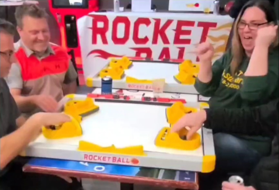Four people playing Rocket Ball arcade game, smiling and enjoying the game, with a sign in the background.