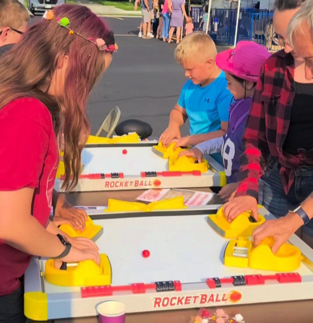 Children and adults playing a tabletop rocket ball game outdoors, focusing on yellow game pieces and a red ball.