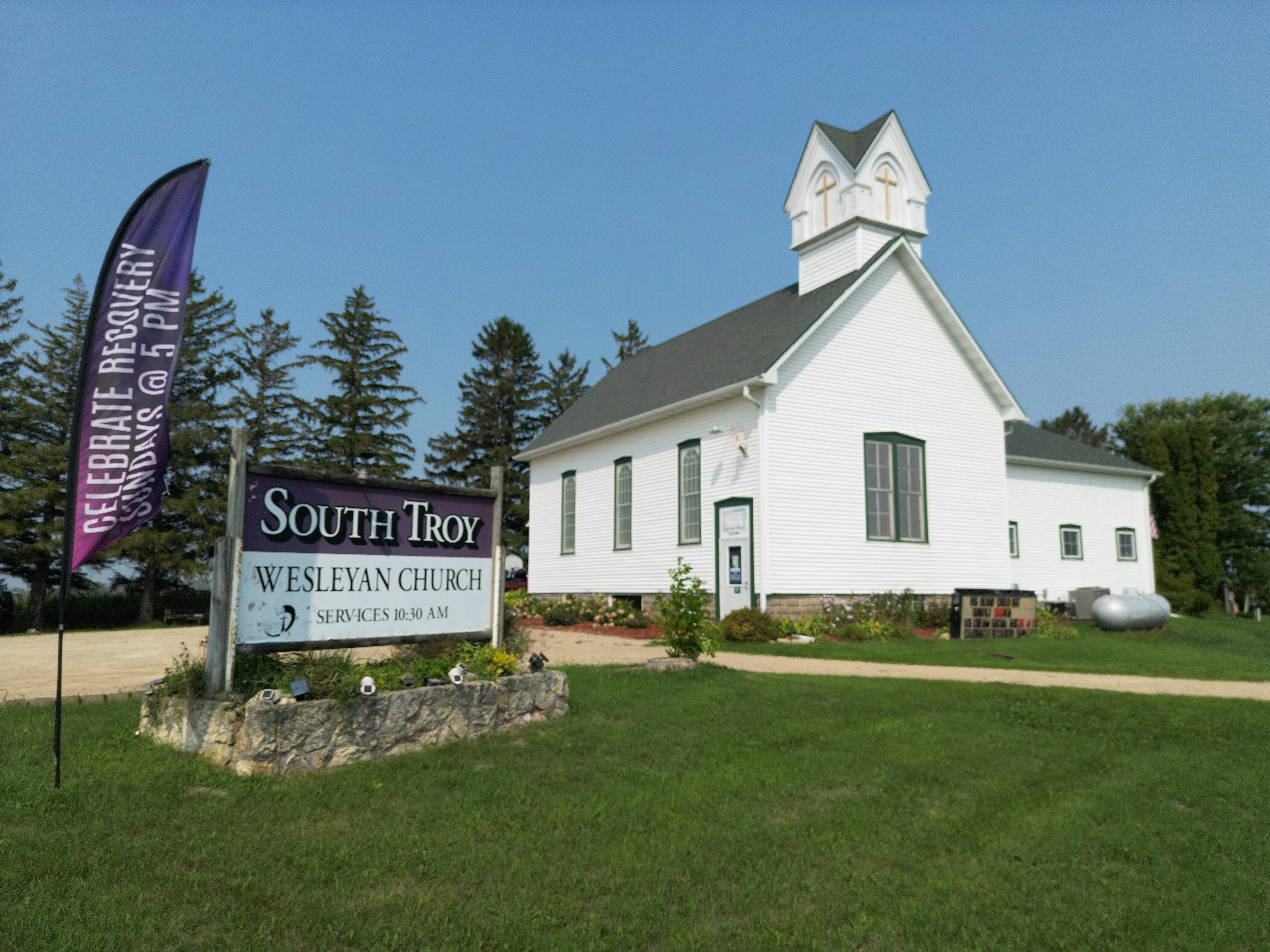 White church with steeple, surrounded by trees and a sign for South Troy Wesleyan Church.