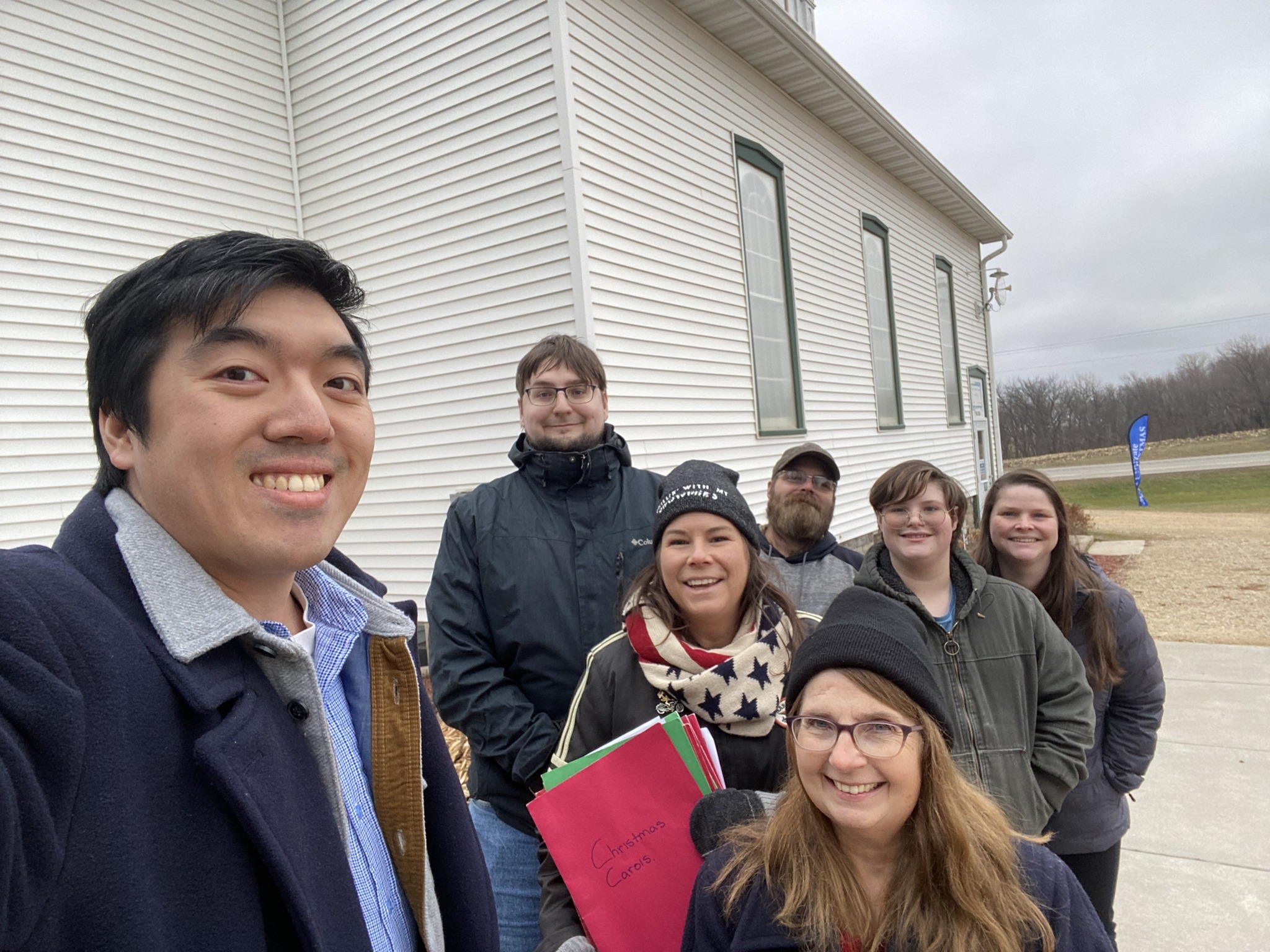 Group of seven people standing outdoors near a white building, smiling, with cloudy sky in background.