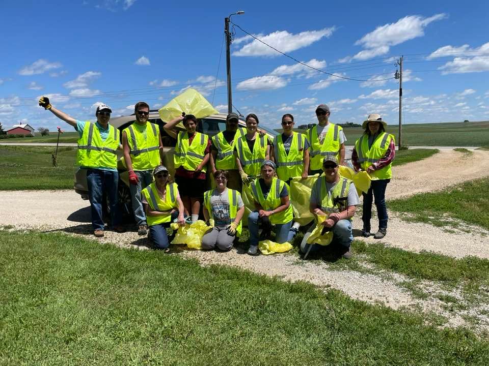 Group of people wearing yellow safety vests standing outdoors on a dirt path with green grass and blue sky.