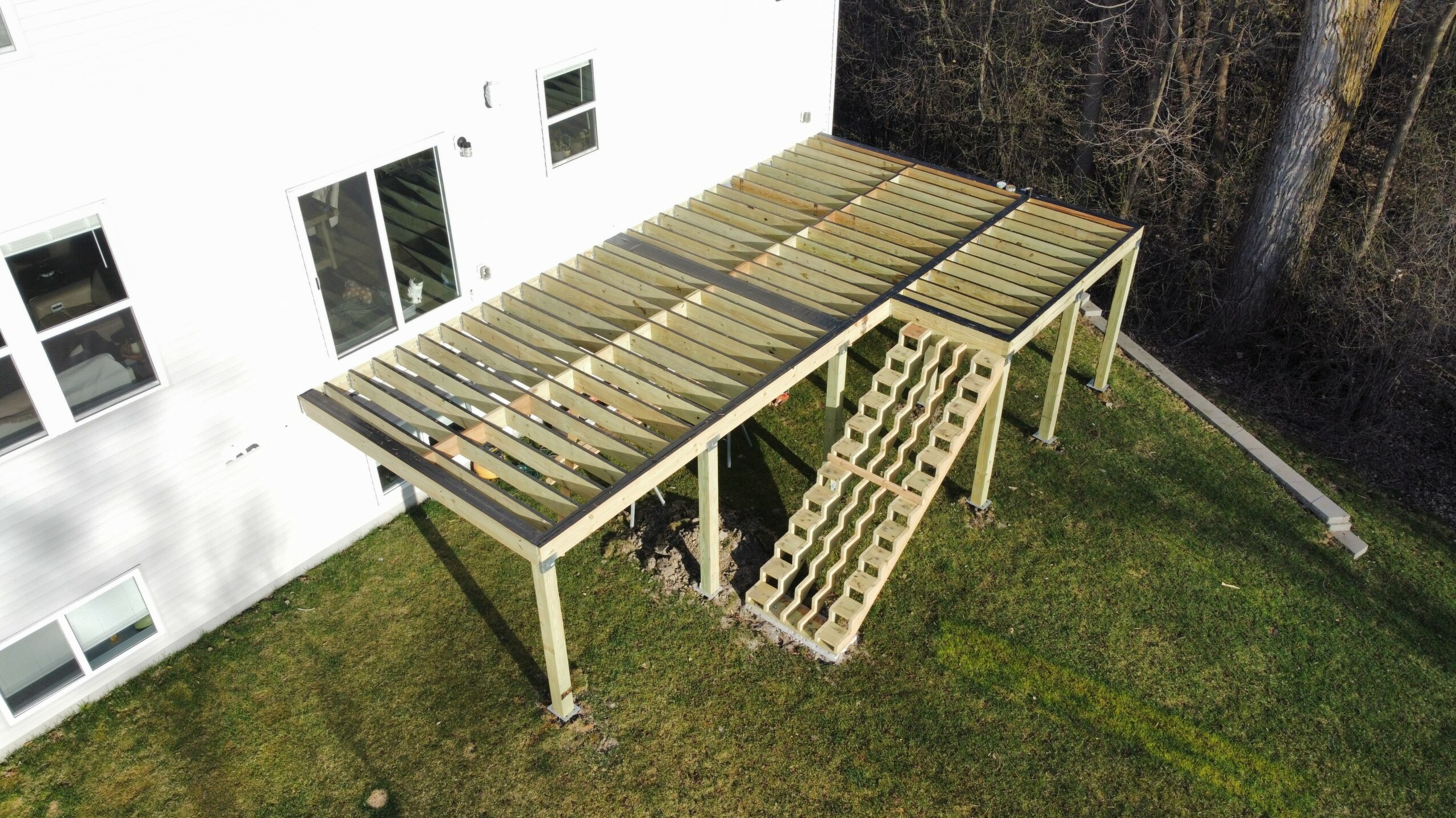 Wooden deck under construction with support beams and a ramp, adjacent to a white building with windows, on grass.