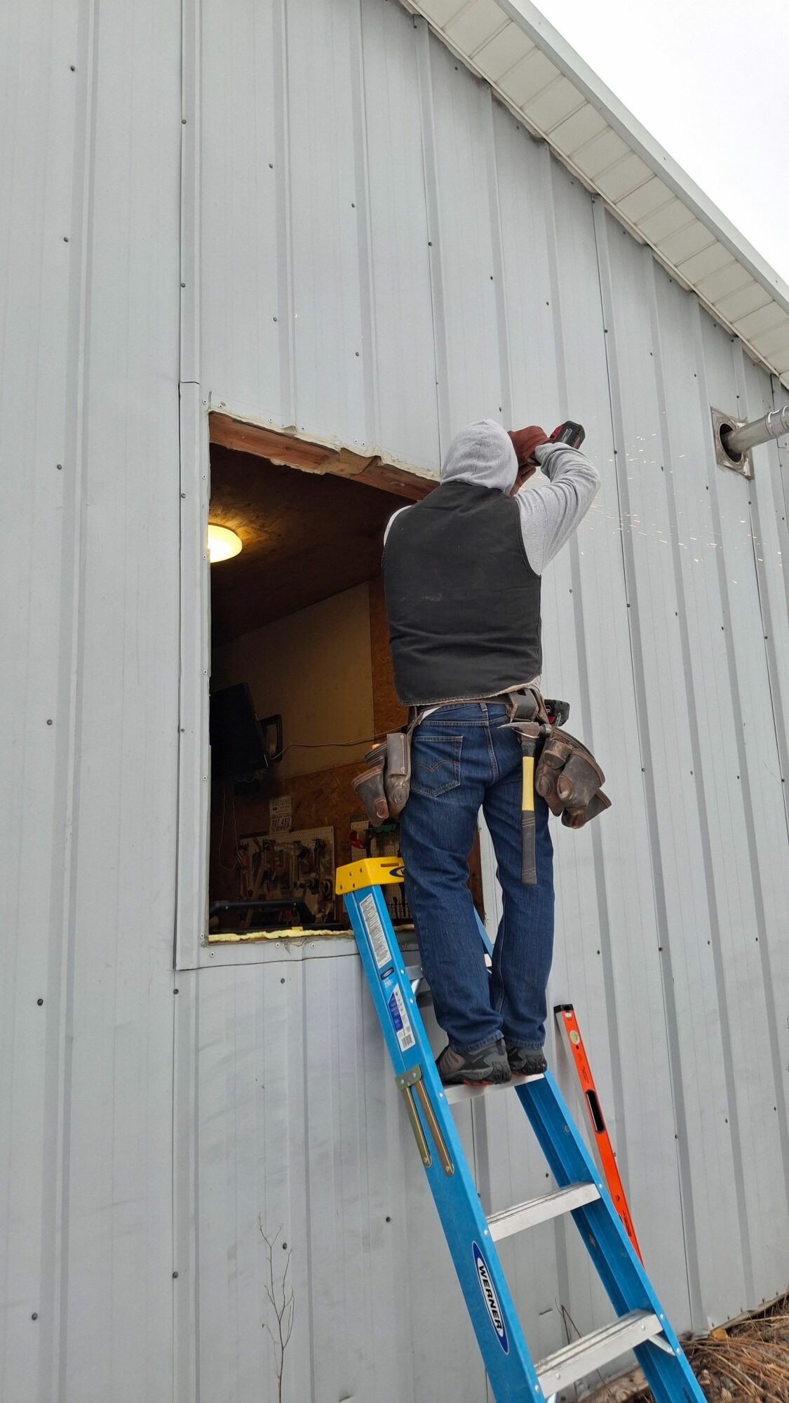 Person standing on a ladder working on a building's exterior wall, window opening, tools hanging from belt.