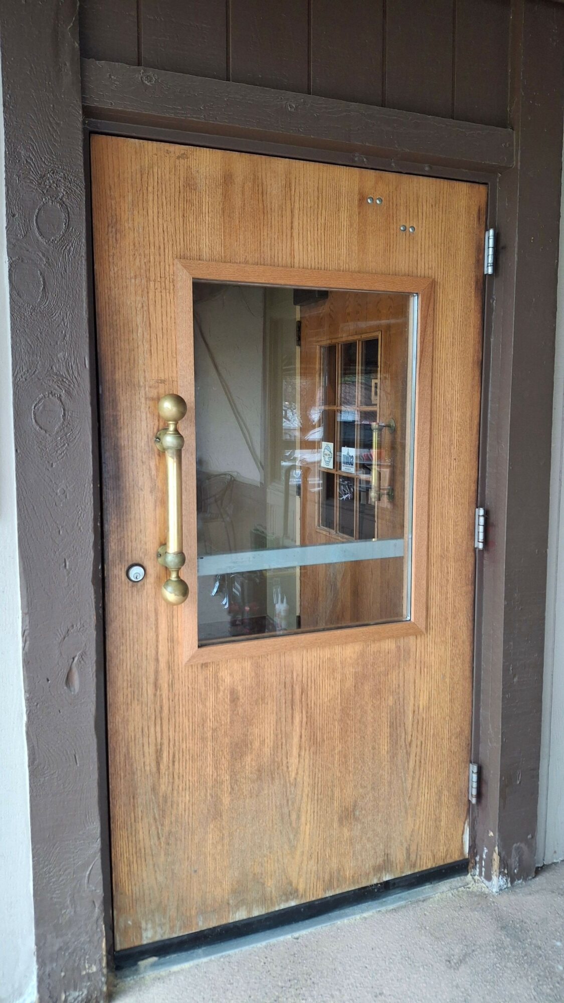 Wooden door with glass window and brass handle, set in a dark frame, with a reflection of a building in the glass.