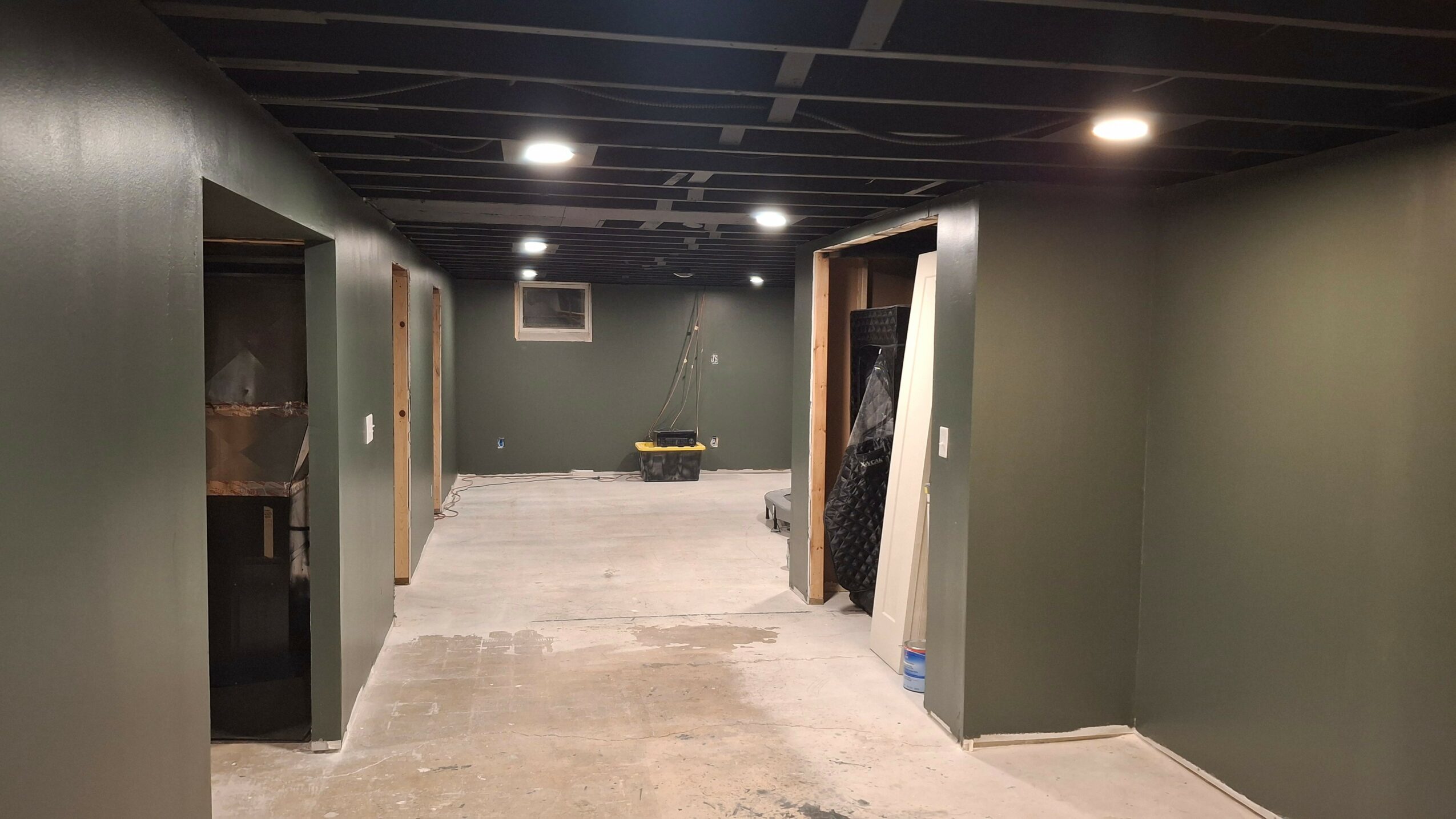 Empty hallway with dark ceiling, green walls, and concrete floor, leading to a small window and doorway at the end.