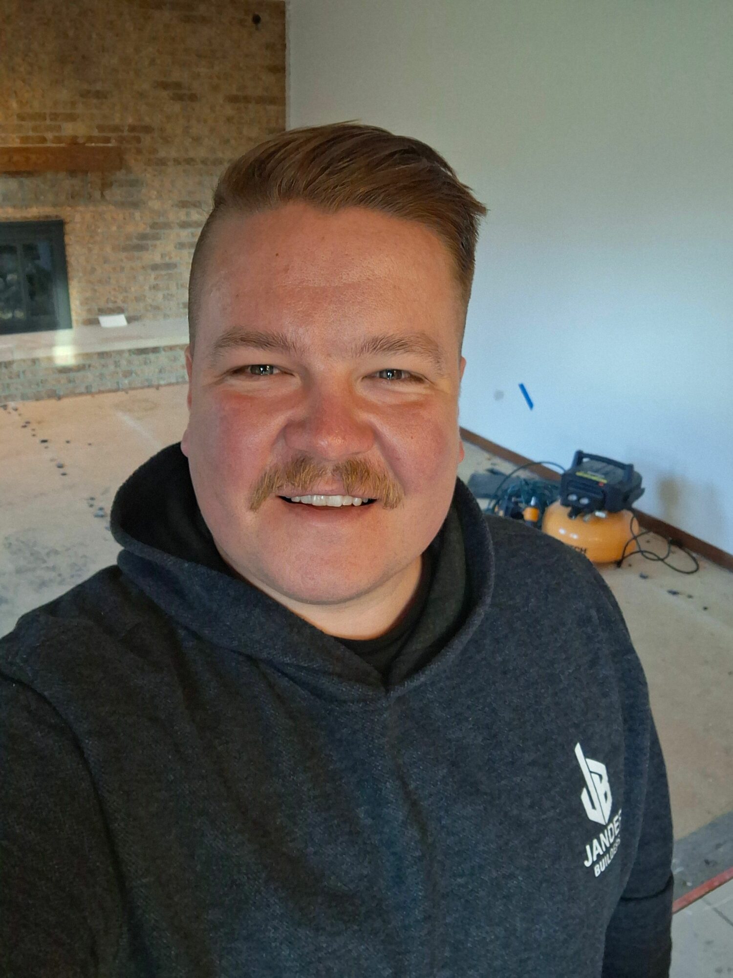 Smiling man with short hair wearing a dark hoodie, indoors with brick wall and equipment in background.