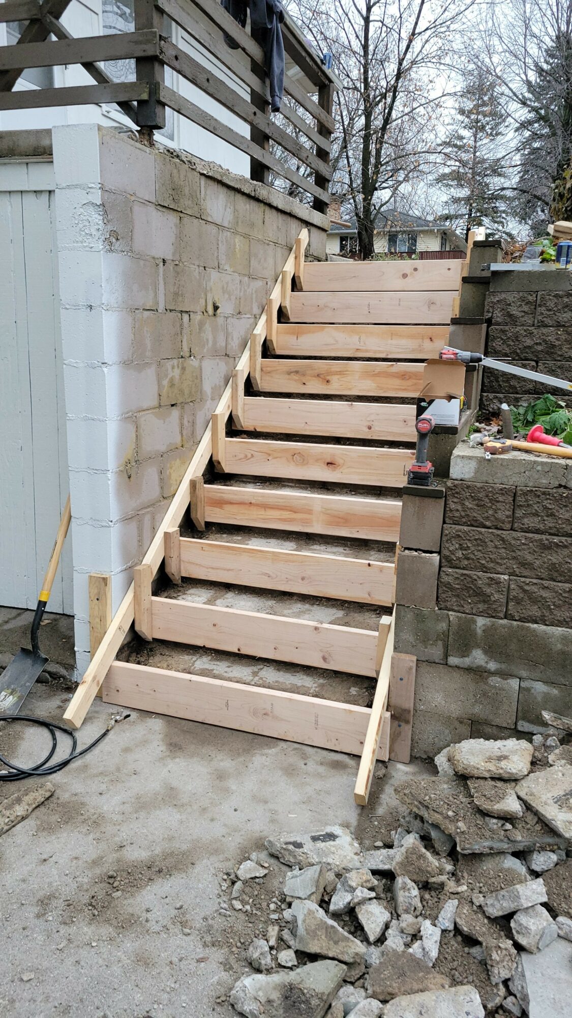 Wooden staircase under construction with steps and support beams, next to a brick wall and outdoor area with trees.