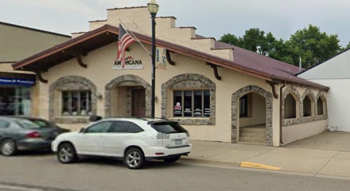 Building with arched windows and a red roof, parked cars in front, American flag on pole, trees in background.