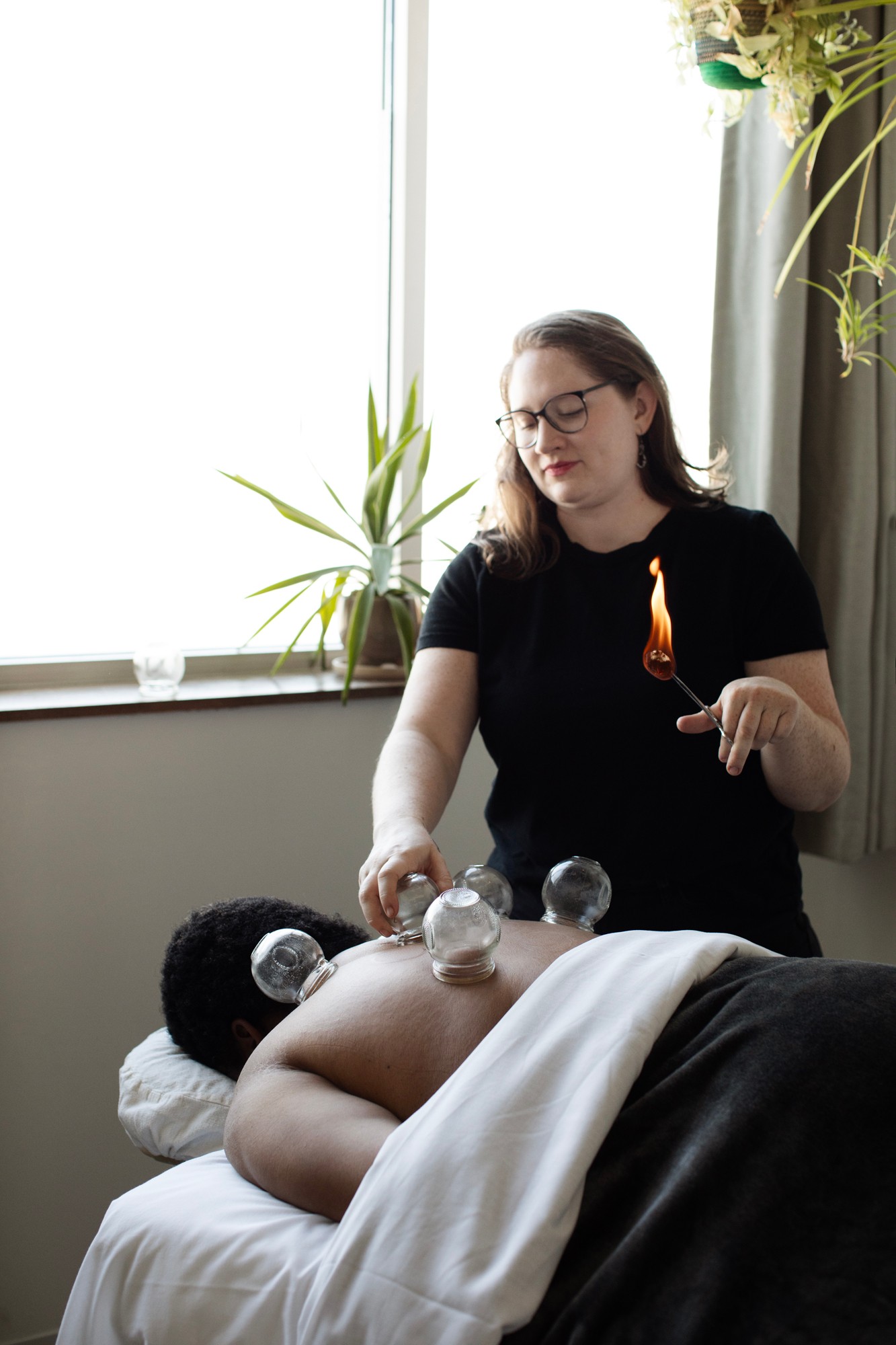Woman with glasses performs cupping therapy on a person's back in a room with a window and plants.