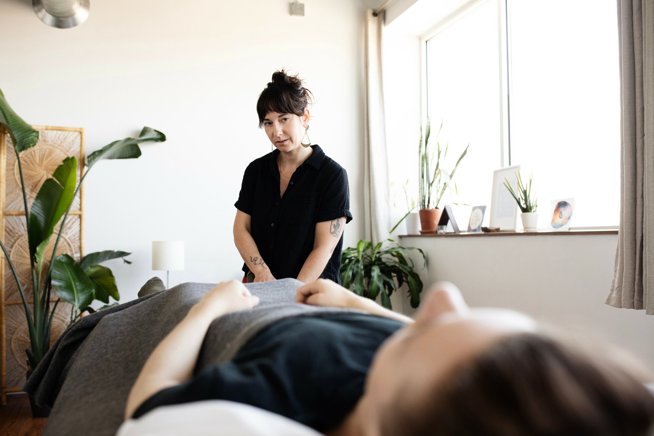 A woman stands beside a person lying on a bed in a bright room with plants and large windows.