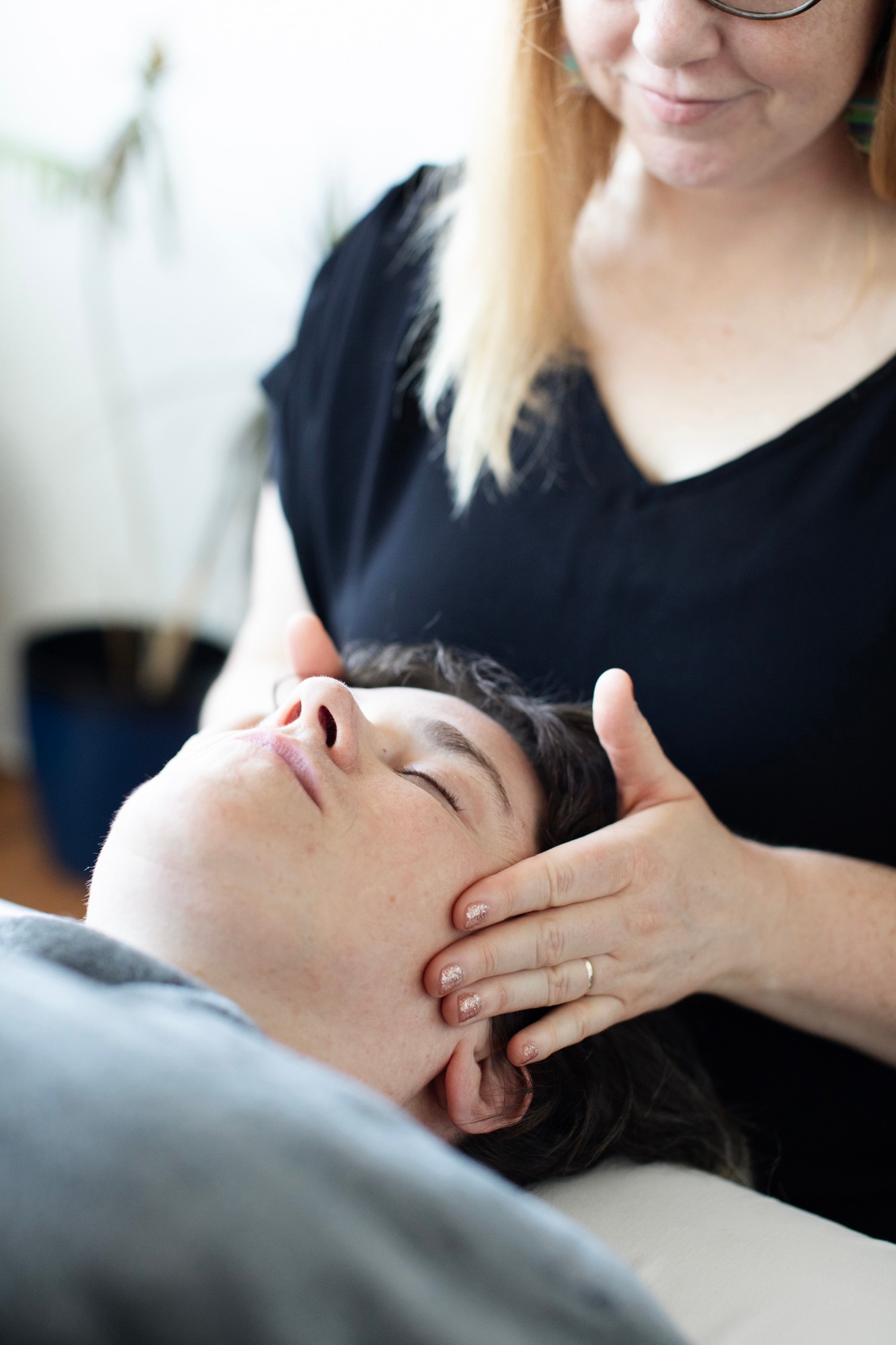 Person receiving a facial massage from a practitioner with glasses and blonde hair.