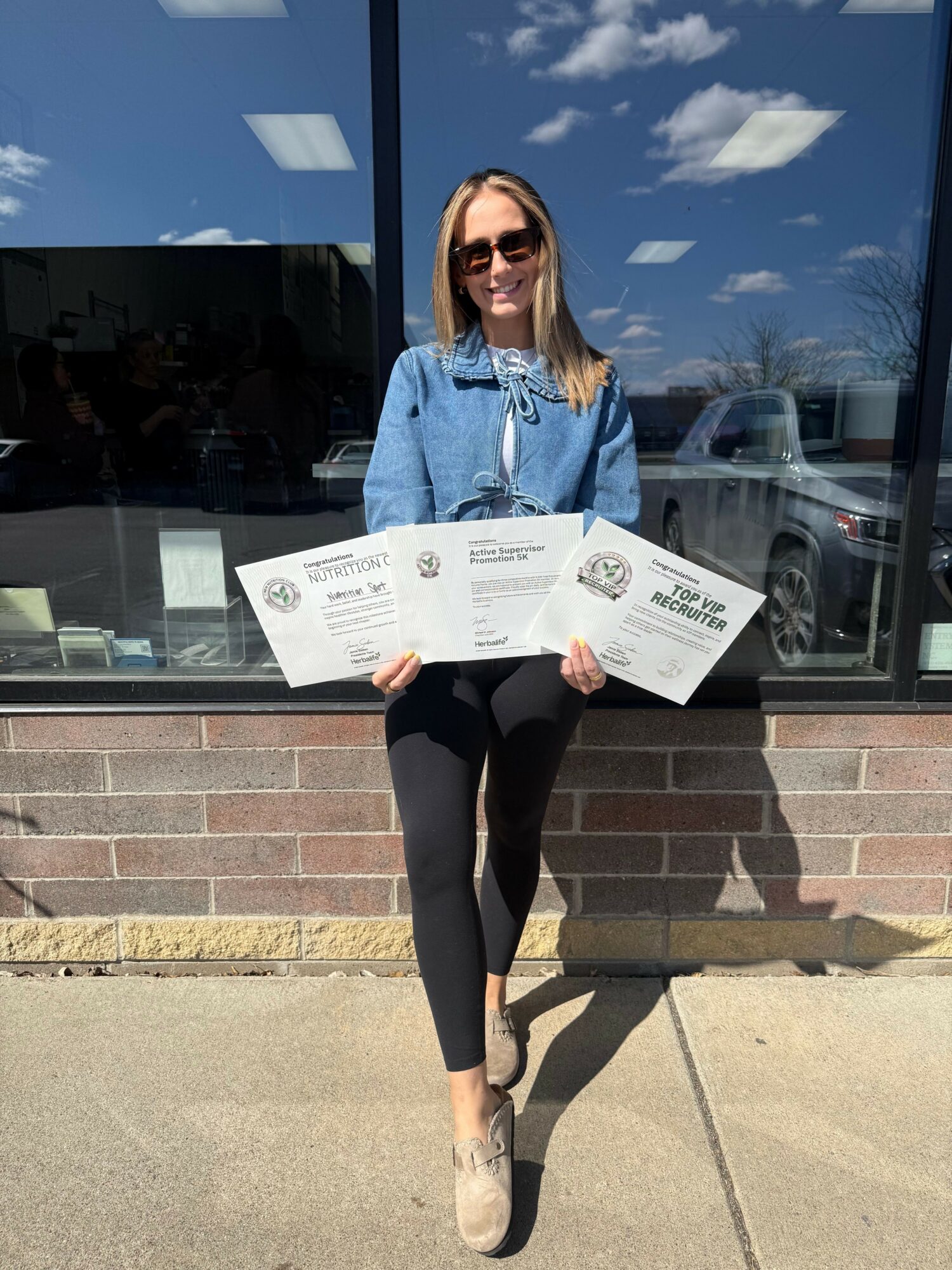 Woman standing outside holding three certificates, wearing sunglasses, denim jacket, black pants, and beige shoes, with a glass window behind her.